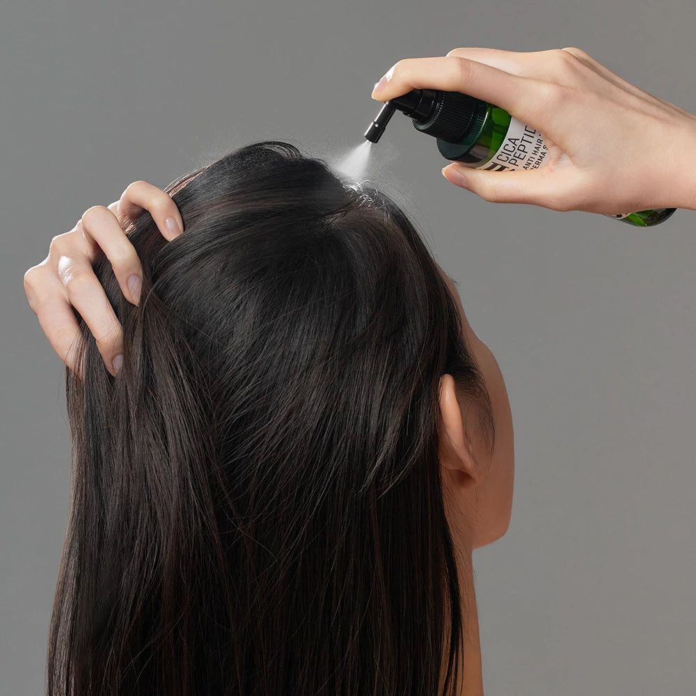 Grüne Flasche mit schwarzem Sprühkopf. Eine Hand sprüht das Produkt auf die Kopfhaut einer Person.