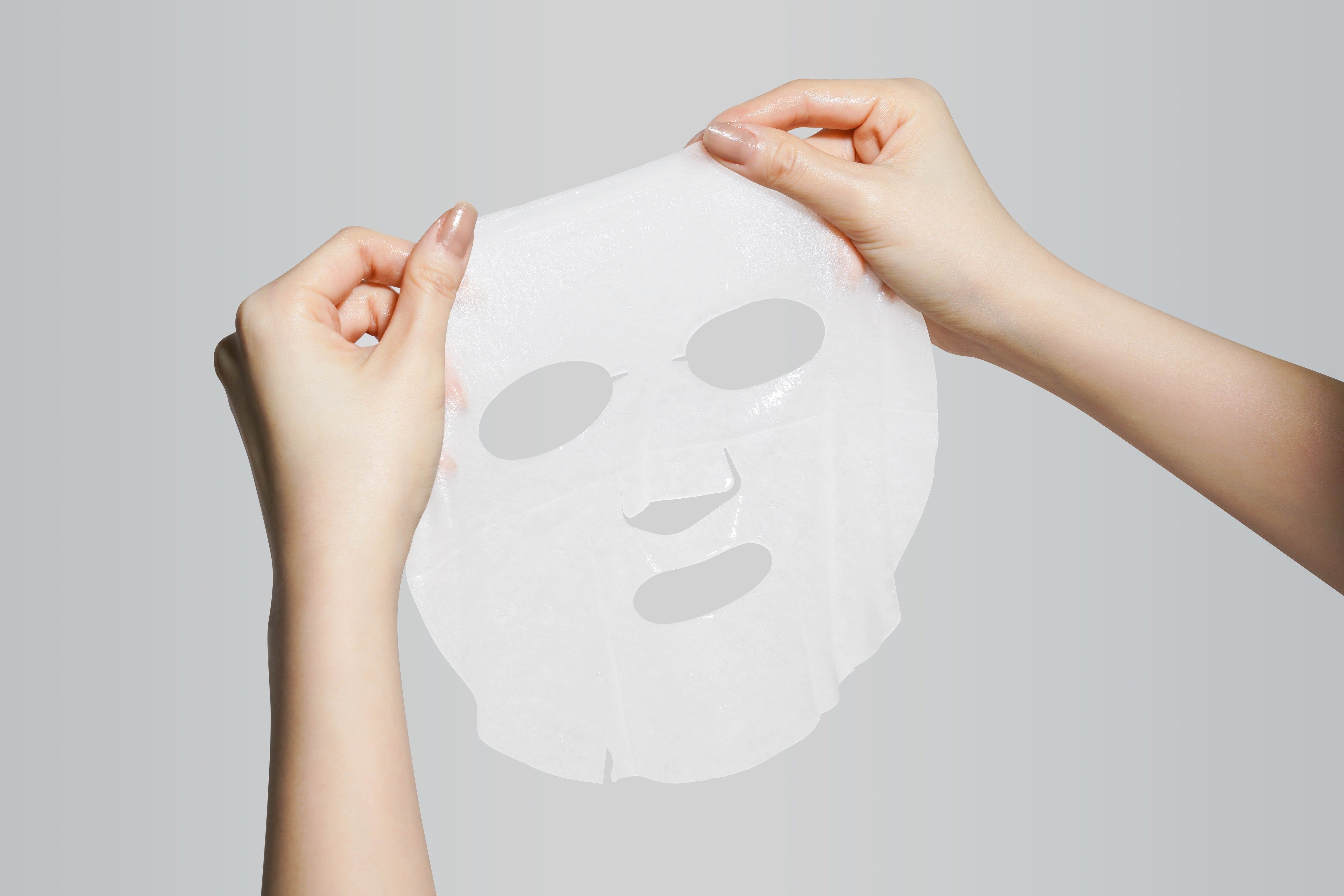 Gesichtsmaske mit Aussparungen für Augen, Nase und Mund. Hände halten Maske vor grauem Hintergrund.