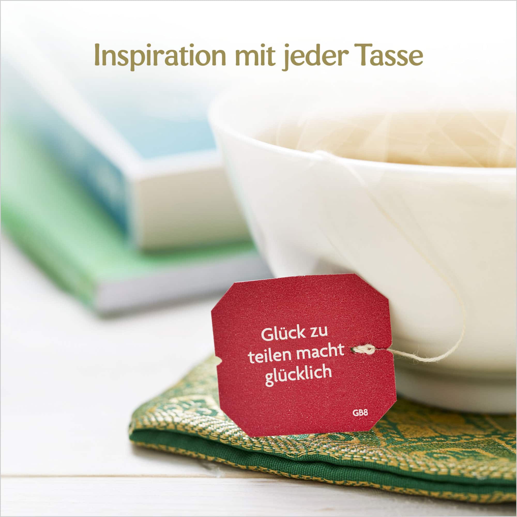 Eine Tasse Tee mit einem roten Teebeutel. Auf dem Teebeutel steht "Glück zu teilen macht glücklich".
