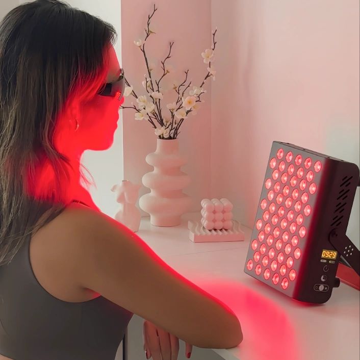 Frau mit Brille vor rotem Lichtgerät. Graues Gerät mit Ständer. Weiße Oberfläche.