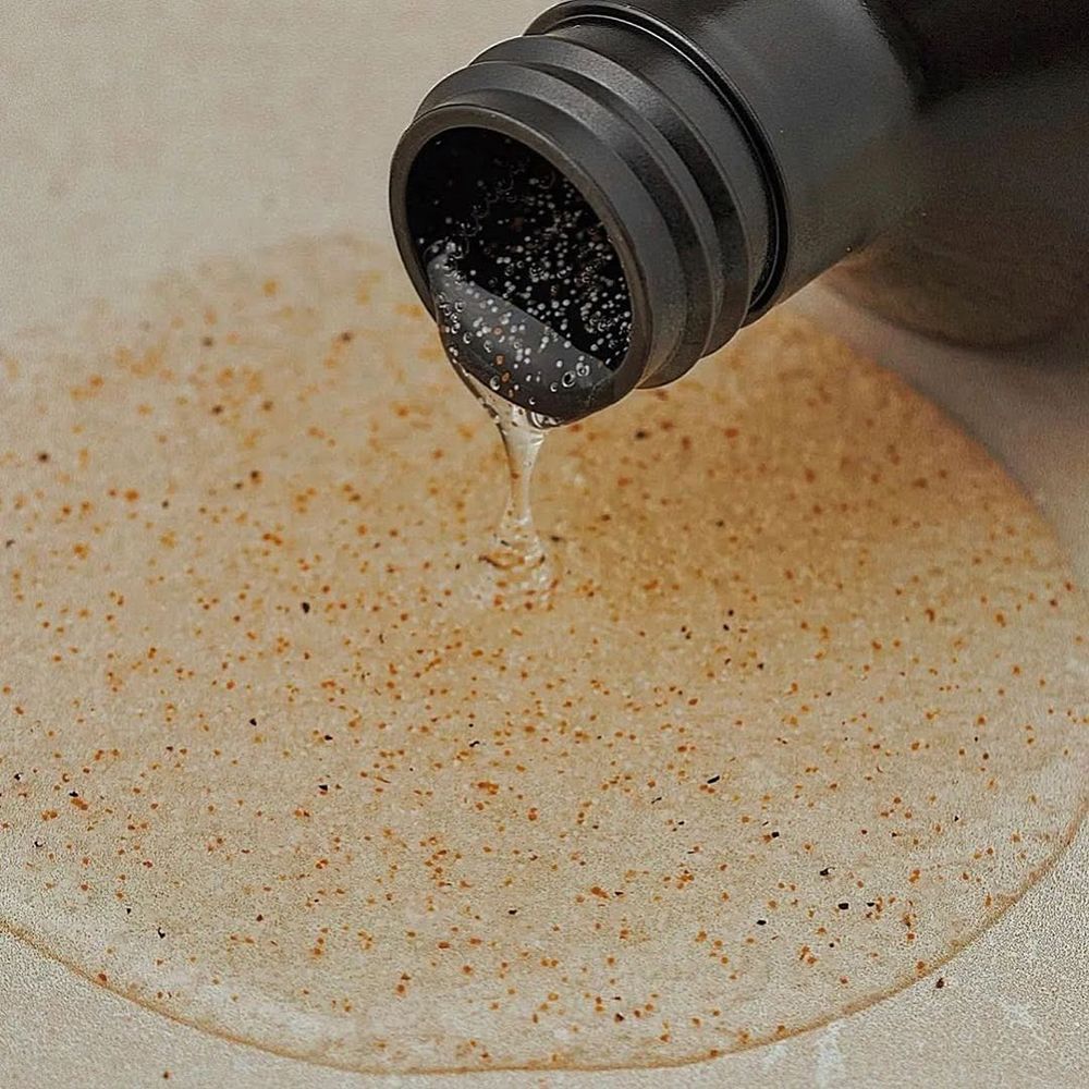 Braune Flasche, aus der Flüssigkeit auf eine Oberfläche tropft. Flüssigkeit enthält Partikel. Schwarzer Pumpverschluss sichtbar.