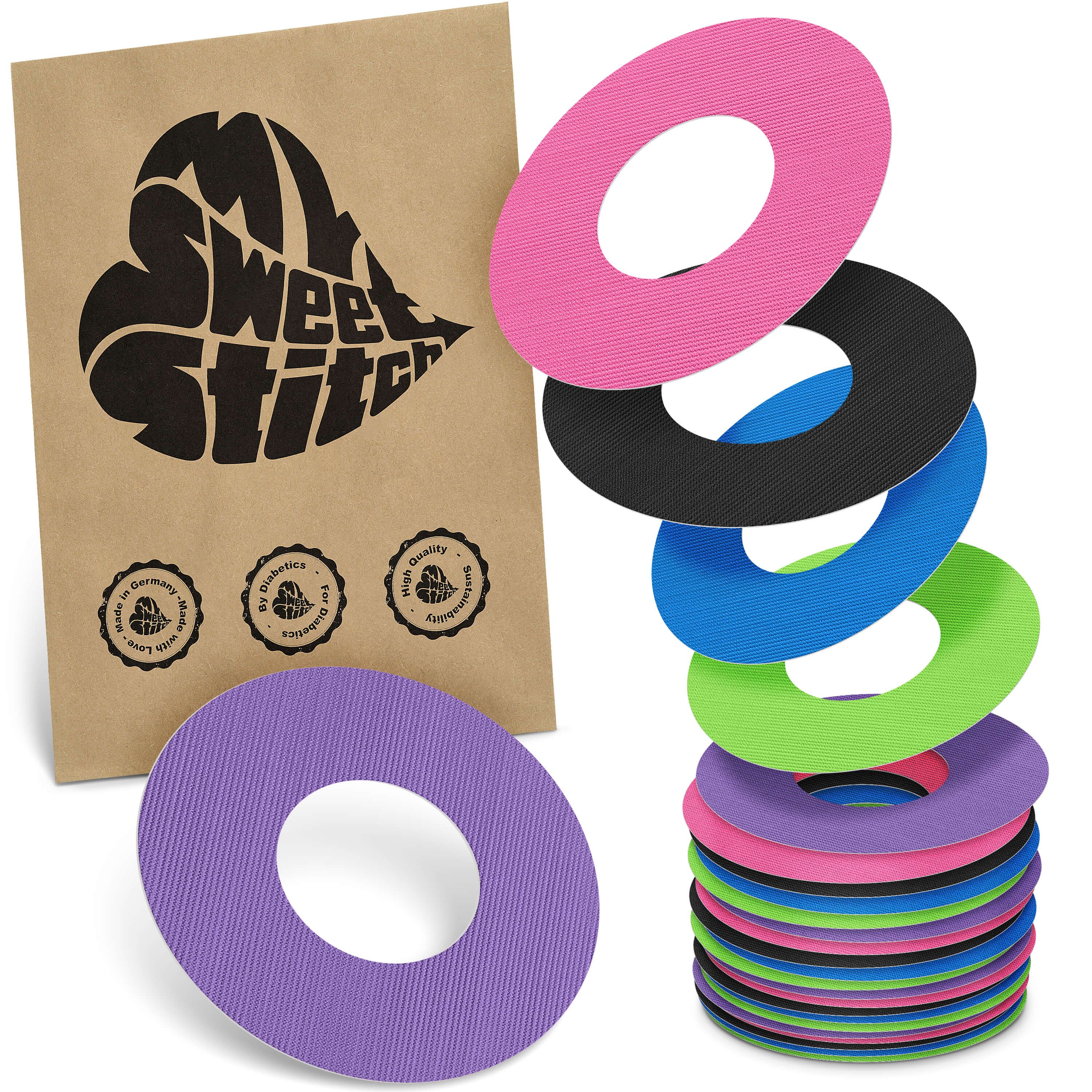 Mehrfarbige Fixierringe, braune Verpackung mit Logo. Ringe in Pink, Schwarz, Blau, Grün, Lila. Stapel und einzelne Ringe sichtbar.