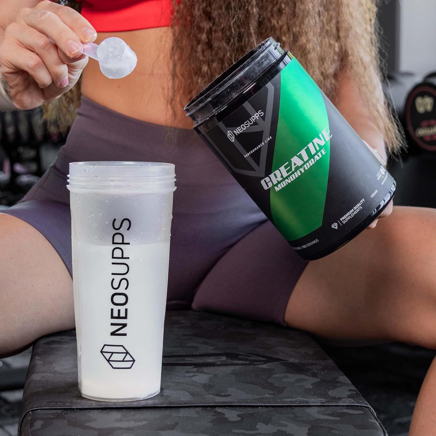Frau gießt Pulver aus Dose in Shaker. Aufschrift: Creatine Monohydrate. Marke: Neosupps. Shaker mit Flüssigkeit.