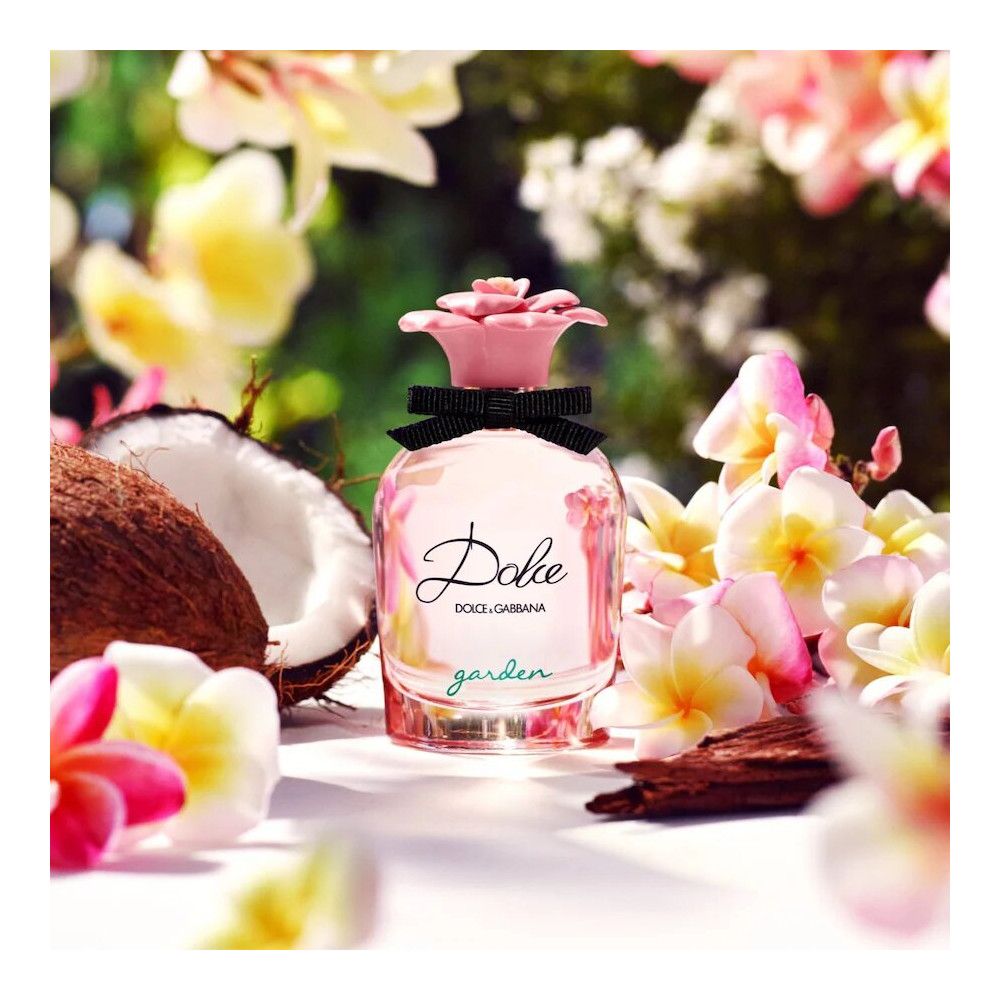 Rosa Parfümflakon mit Blütenkappe und schwarzer Schleife. Aufschrift "Dolce" und "Dolce & Gabbana". Umgeben von Kokosnuss und Blumen.