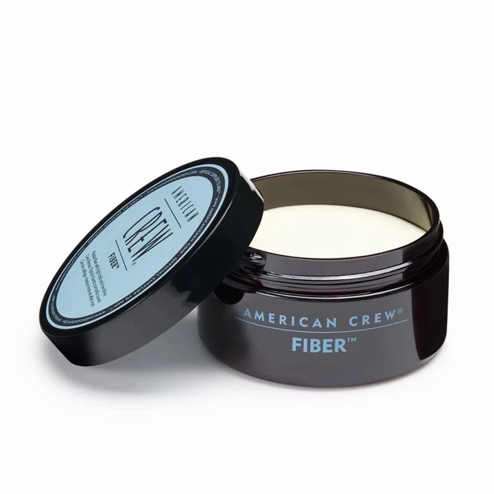 Geöffnete Dose mit Deckel daneben. Aufschrift: AMERICAN CREW FIBER. Produkt sichtbar. Schwarze Dose, blauer Deckel.