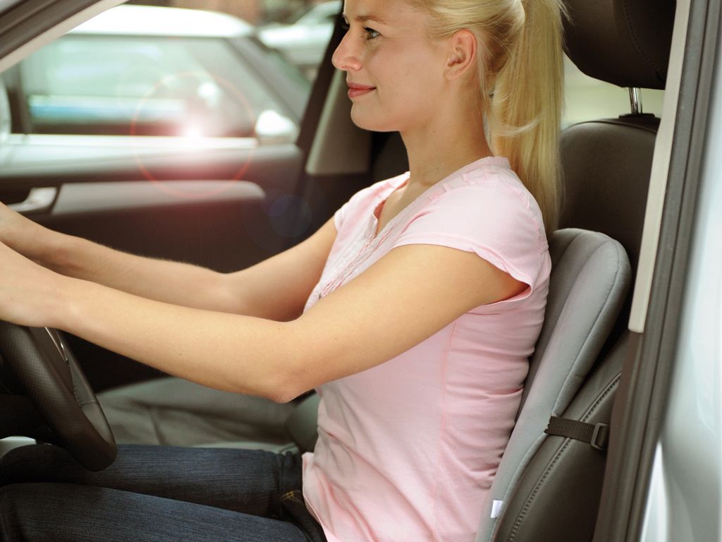 Frau im Auto, die ein graues Rückenkissen benutzt. Sie sitzt am Steuer und lächelt.