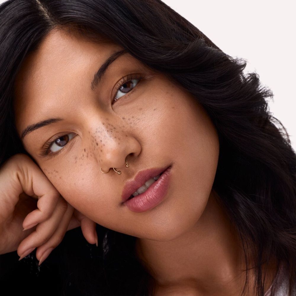 Porträt einer Frau mit dunklen Haaren. Sie stützt ihr Kinn auf ihre Hand. Sie hat Sommersprossen und trägt Lippenstift.