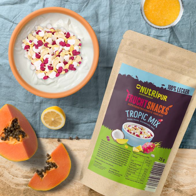 Beutel mit gefriergetrockneten Früchten Tropic-Mix neben Schale mit Joghurt und Früchten. Papaya und Zitrone daneben.