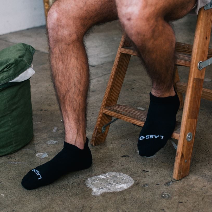Person sitzt auf einer Holzleiter, trägt schwarze Socken mit Lasso-Logo. Füße im Fokus.