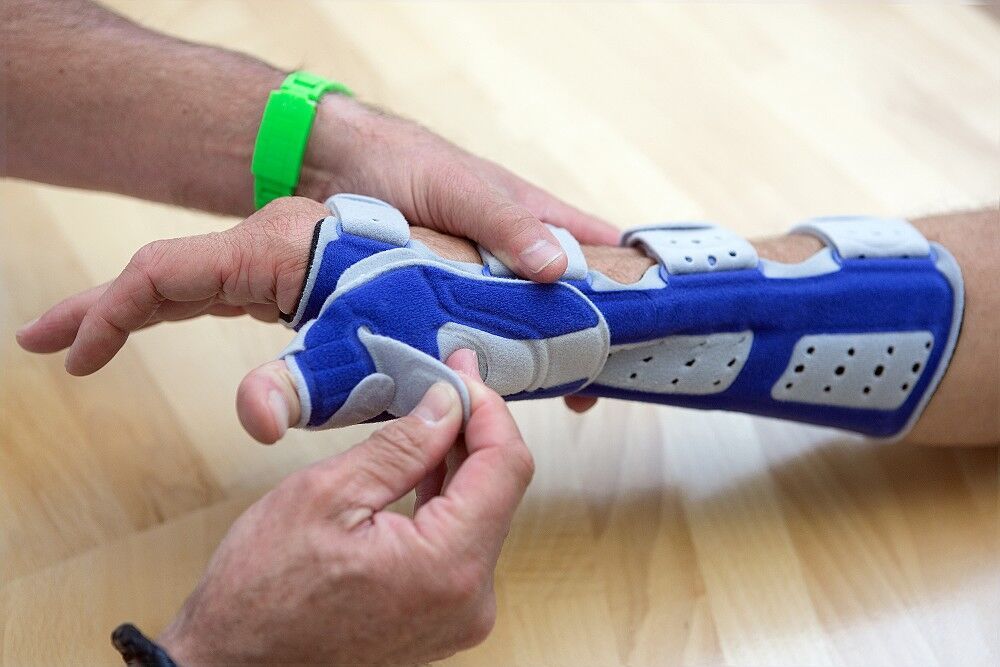 Handgelenk-Daumenorthese ManuLoc Rhizo, blau-grau, am Handgelenk einer Person. Eine andere Person justiert die Orthese.