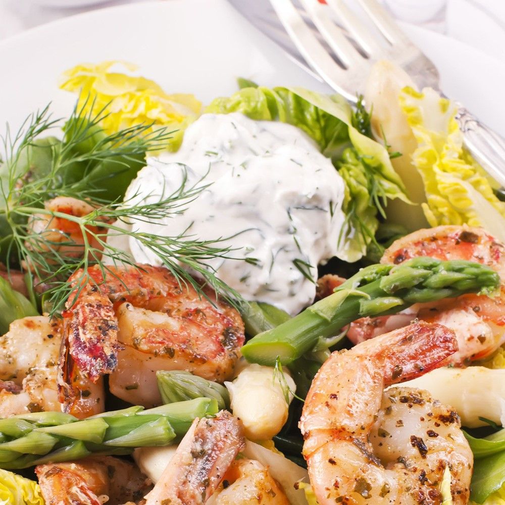 Teller mit Garnelen, Spargel, Salat und Sauce. Garnelen gebraten. Grüner Spargel. Salatblätter. Weiße Sauce.