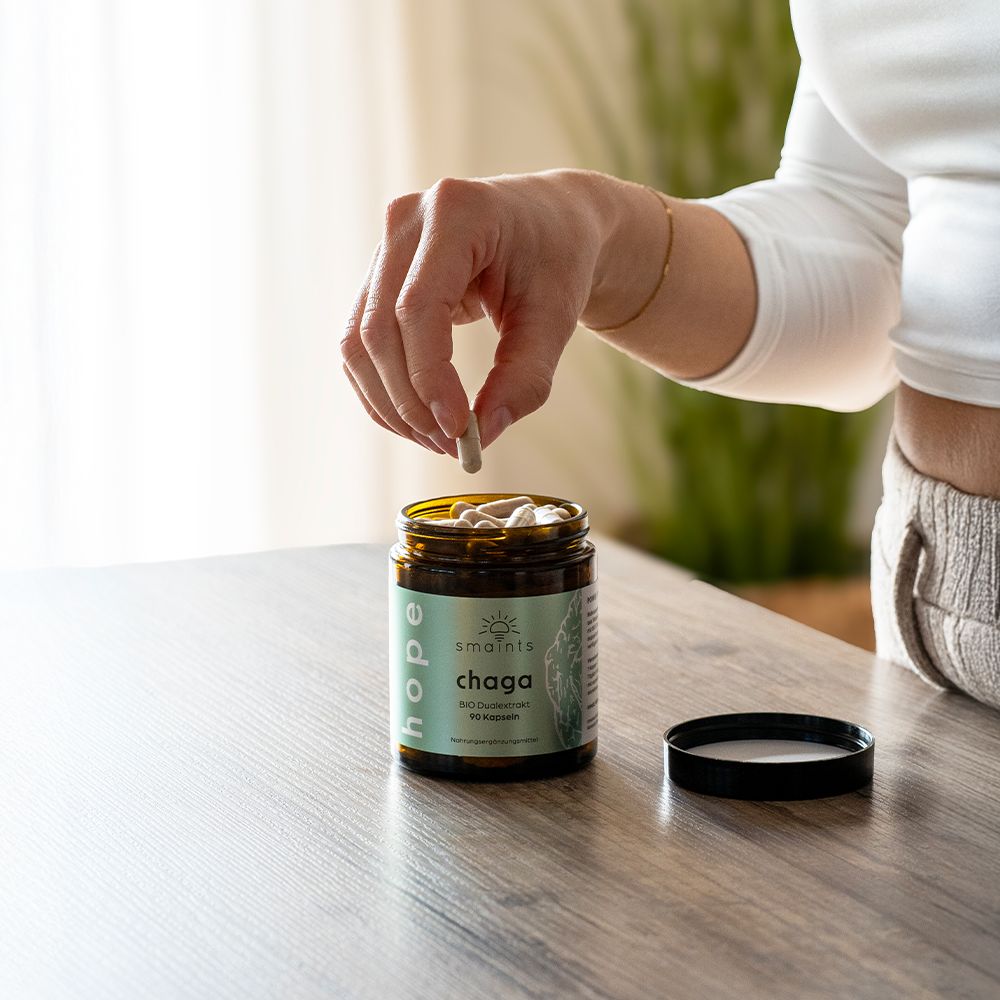 Person hält Kapsel über offener Dose. Dose mit Kapseln auf Tisch. Schwarzer Deckel daneben.