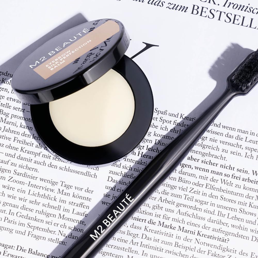 Offener Tiegel M2Beauté Eyebrow Silk Perfection Balm und Bürste. Aufschrift: M2 BEAUTÉ. Produkt auf Zeitung. Creme-farbenes Produkt sichtbar.