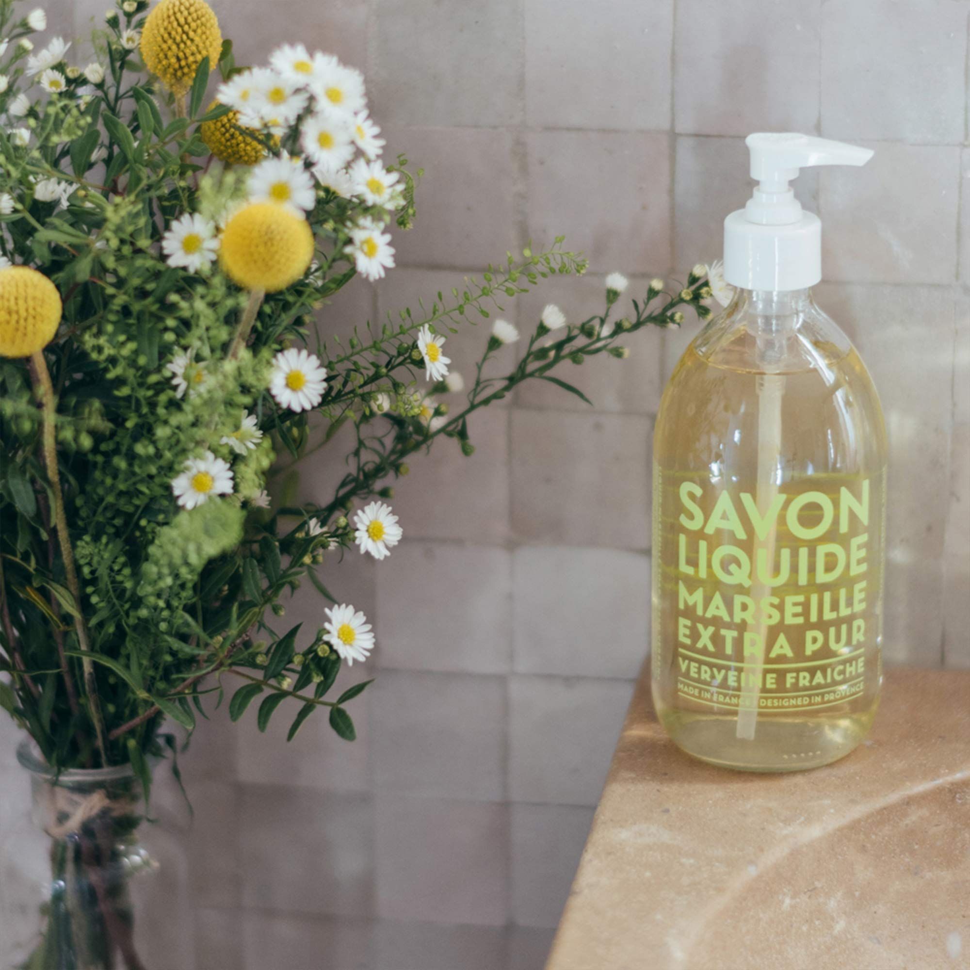 Transparente Flasche mit weißem Pumpkopf neben Blumenstrauß. Aufschrift: Savon Liquide Marseille Extra Pur, Verveine Fraîche.