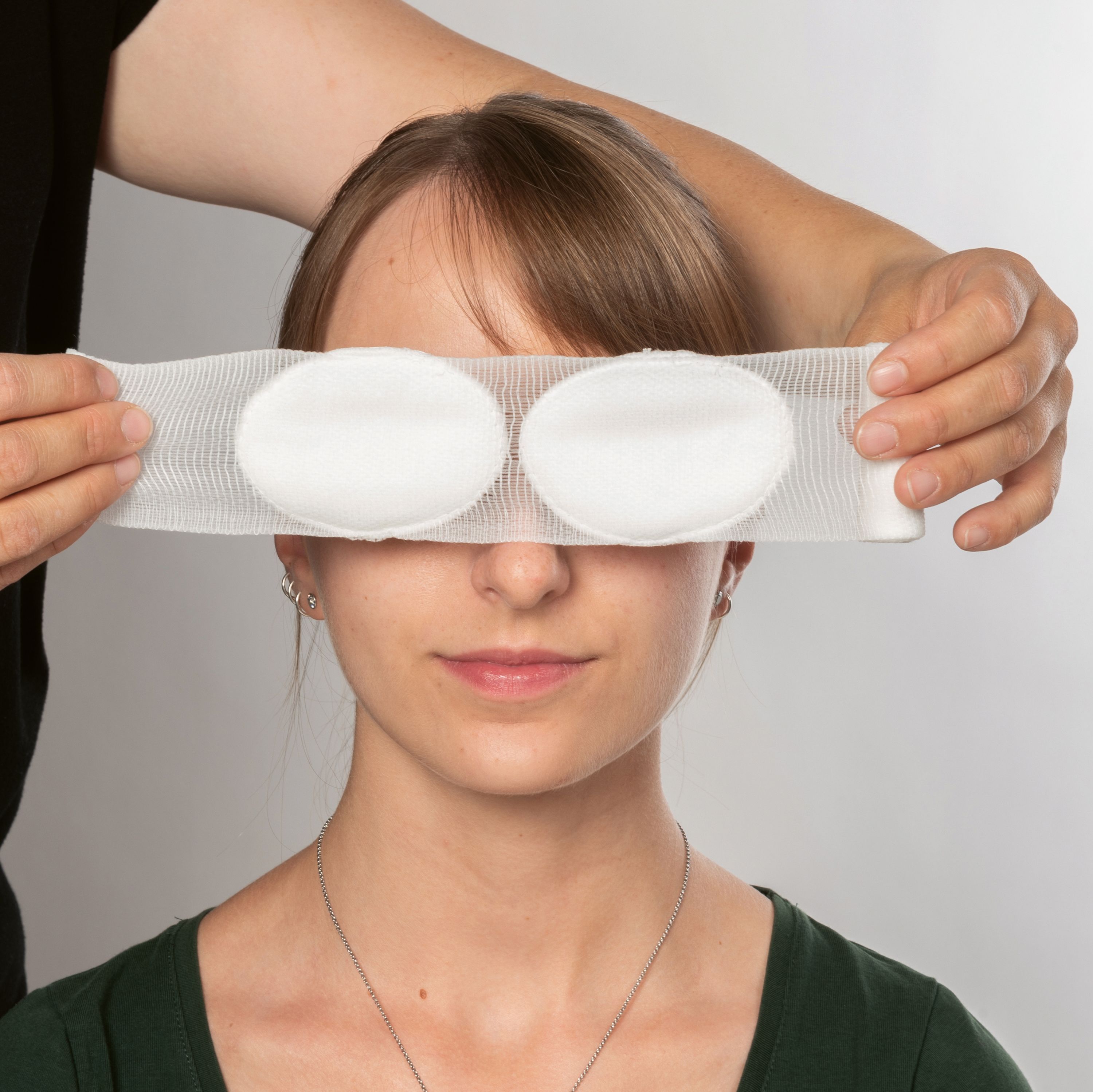 Person mit Augenverband. Zwei weiße Kompressen in einem Netzverband vor den Augen. Hände halten den Verband.