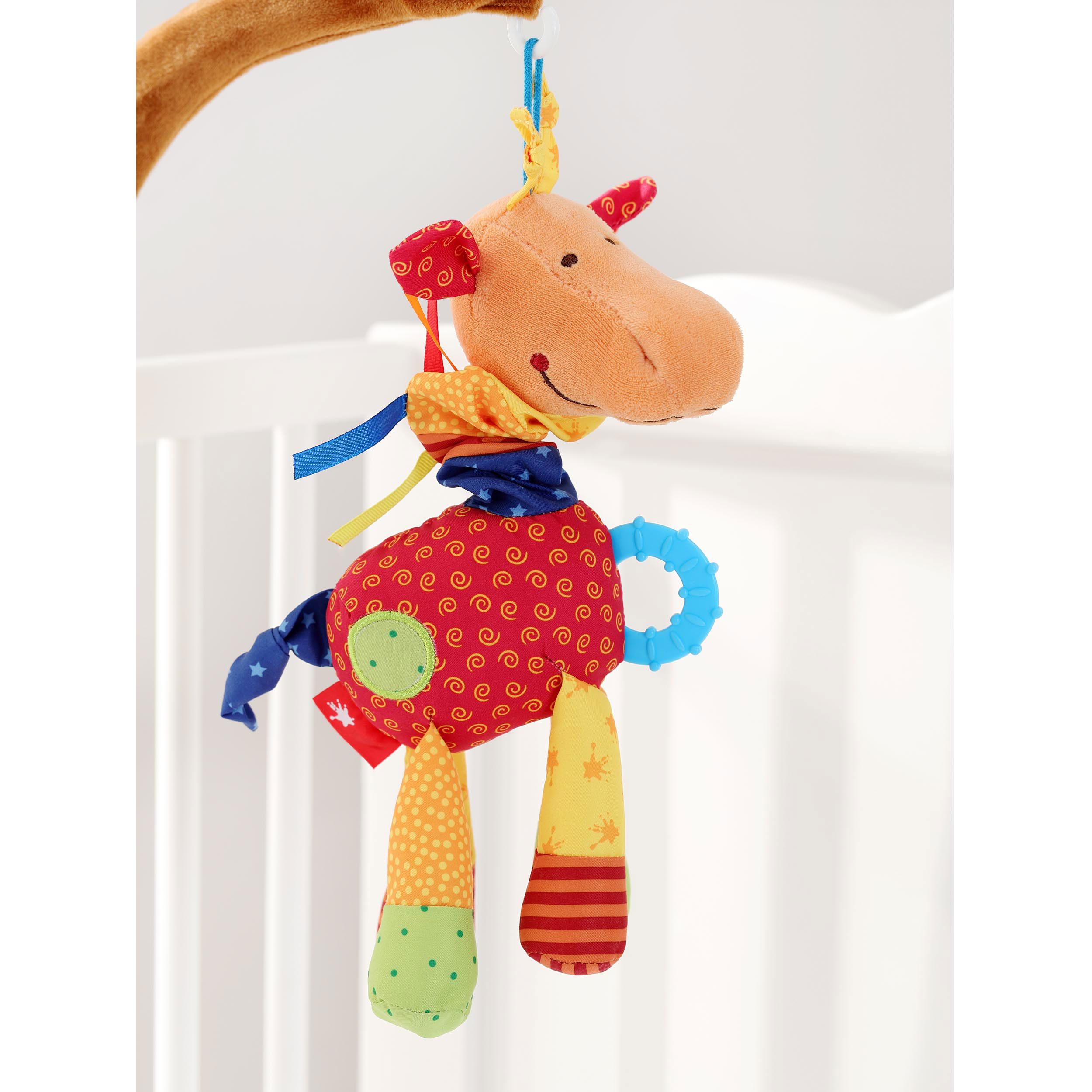 Spielfigur hängt an einem Babybett. Orangefarbenes Tier mit bunten Stoffteilen. Blaue Ringe, gelbe und rote Akzente.