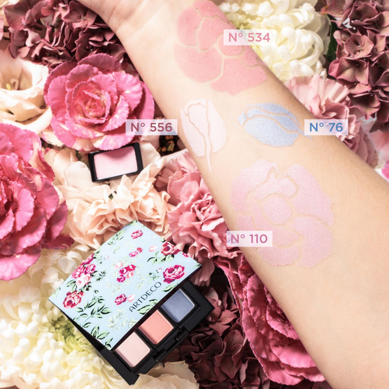 Arm mit verschiedenen Lidschatten-Farben. Artdeco-Palette mit vier Farben. Blumen im Hintergrund. Farbnummern auf dem Arm.