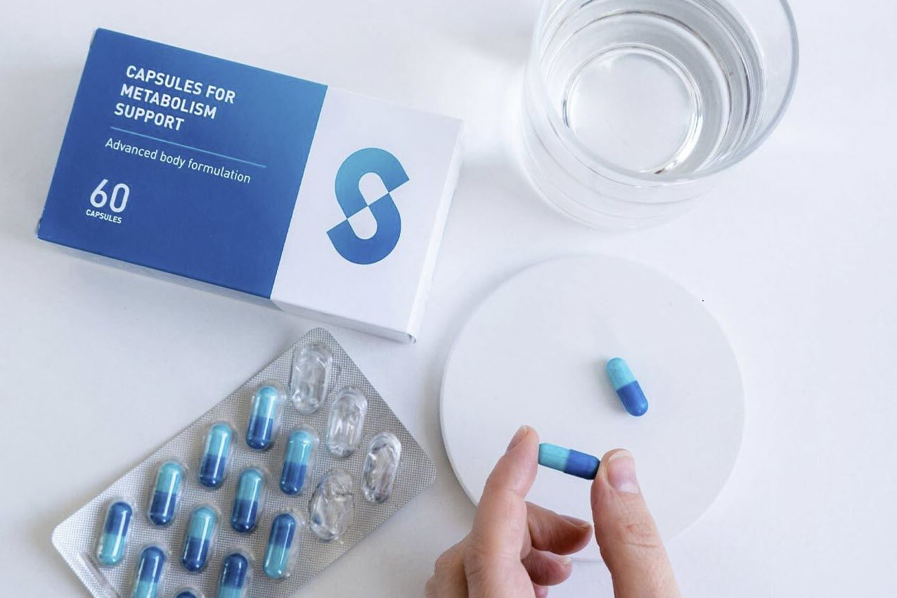 Blau-weiße Schachtel mit "CAPSULES FOR METABOLISM SUPPORT" neben Kapseln und Glas Wasser. Eine Kapsel wird gehalten.