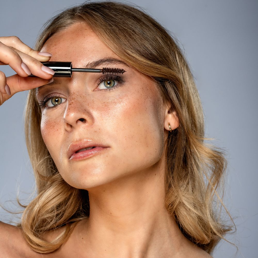 Frau trägt braunen Mascara auf. Schwarzer Stiel, braune Bürste.