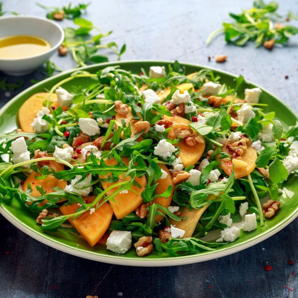Salat mit Kaki, Rucola, Feta und Walnüssen. Auf einem grünen Teller. Mit Dressing in einer weißen Schale.