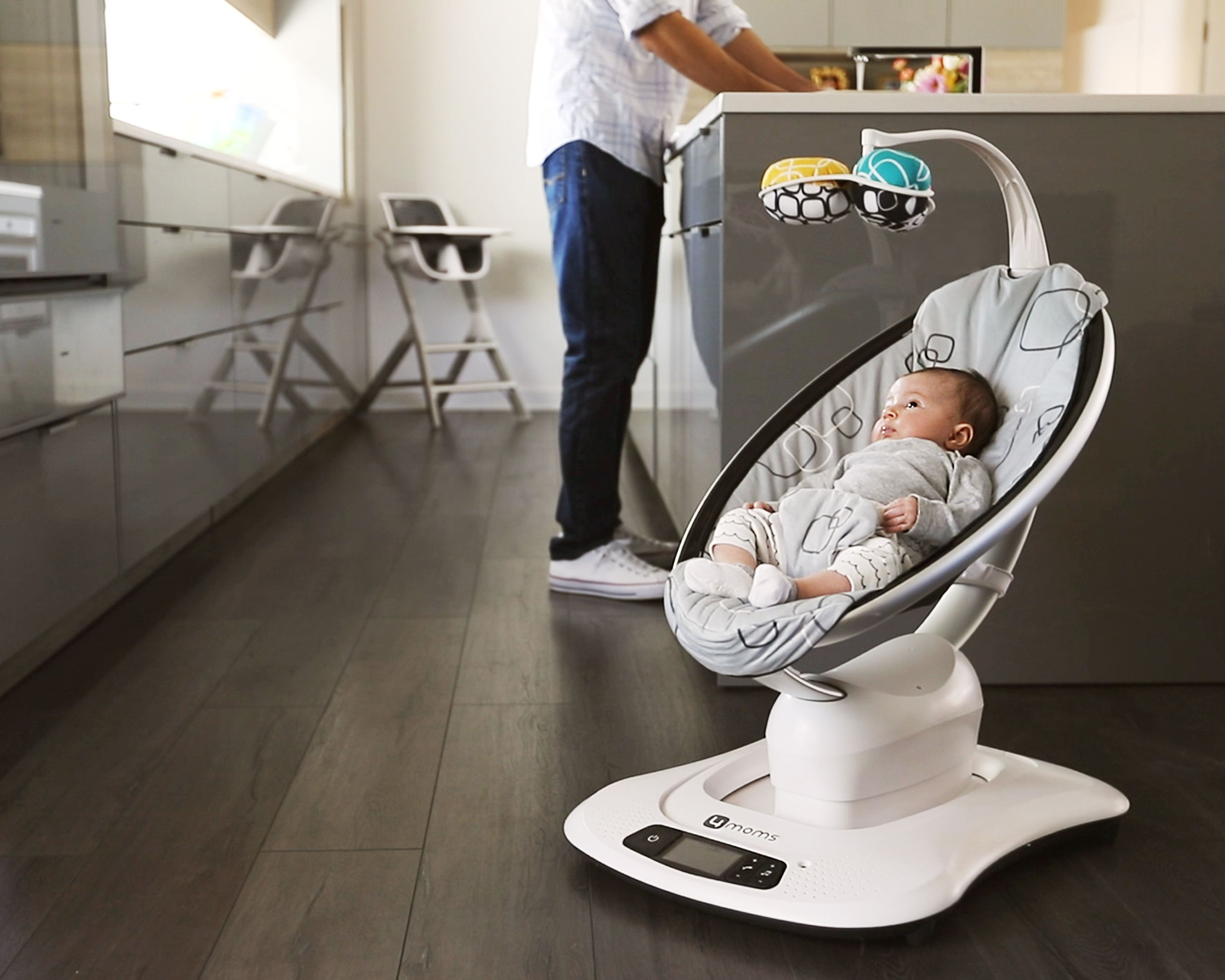 Baby in Babywippe. Grauer Sitz mit Muster, Spielzeug. Person im Hintergrund. Innenraum.