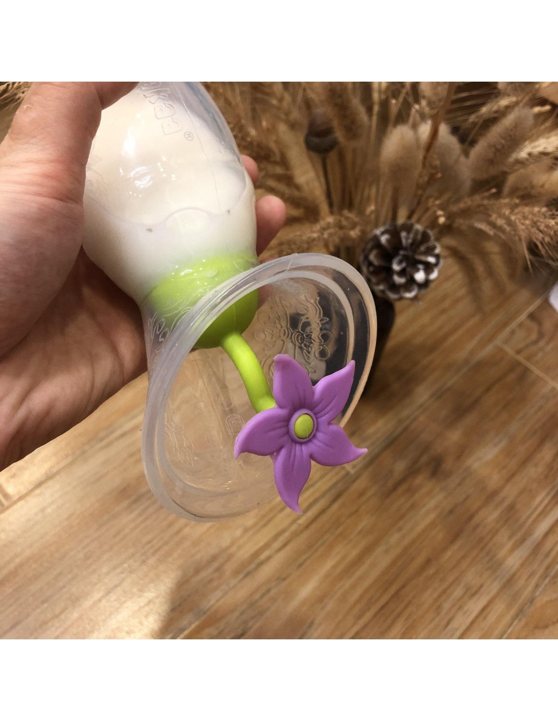 Transparente Milchpumpe mit grünem Verschluss und lila Blütenverschluss. Hand hält die Pumpe.