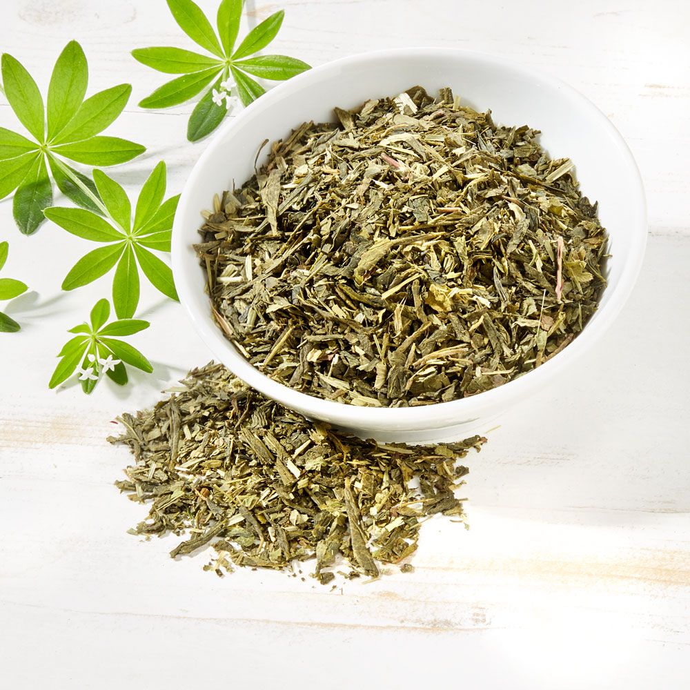 Grüner Tee mit Waldmeister in weißer Schale. Einige Blätter liegen daneben. Grüner Tee und Blätter auf Holzgrund.