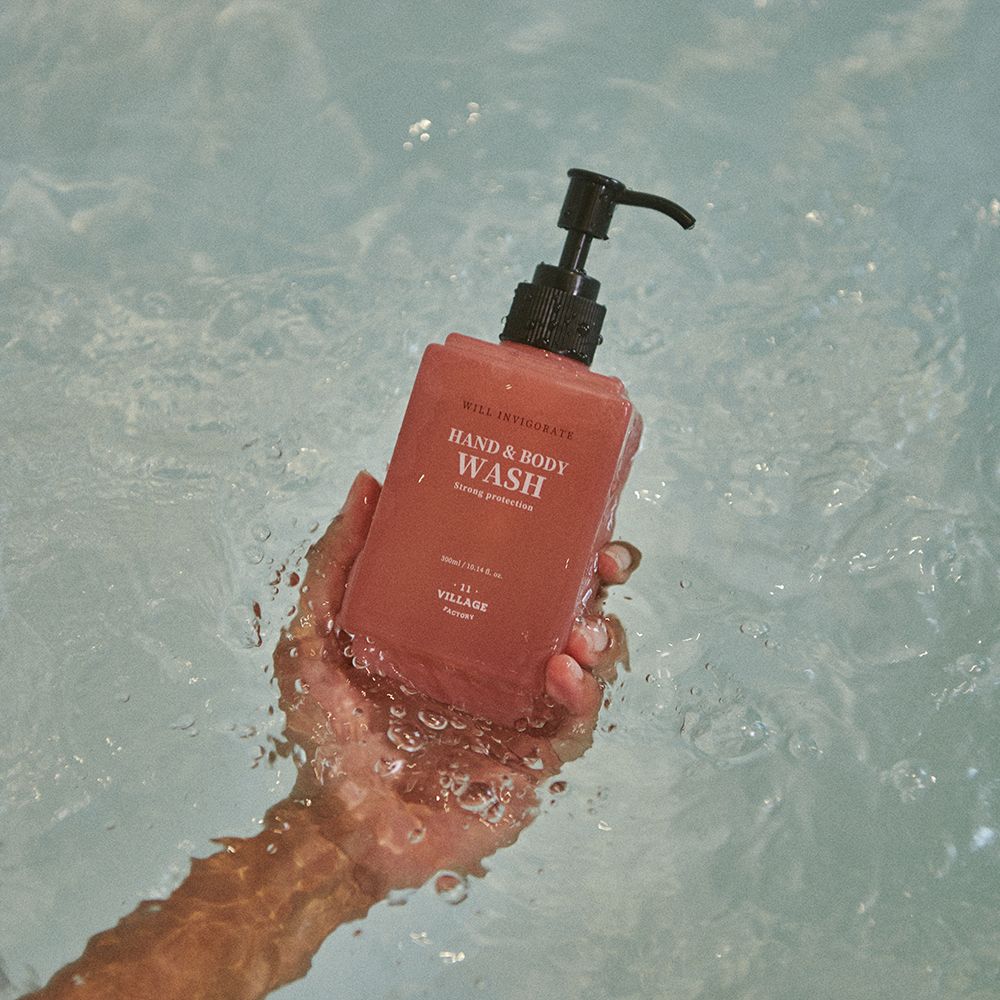 Hand hält rosa Hand- und Körperwaschmittel-Flasche in Wasser. Schwarzer Spender. Aufschrift: Hand & Body Wash, Strong protection, 11 Village Factory.