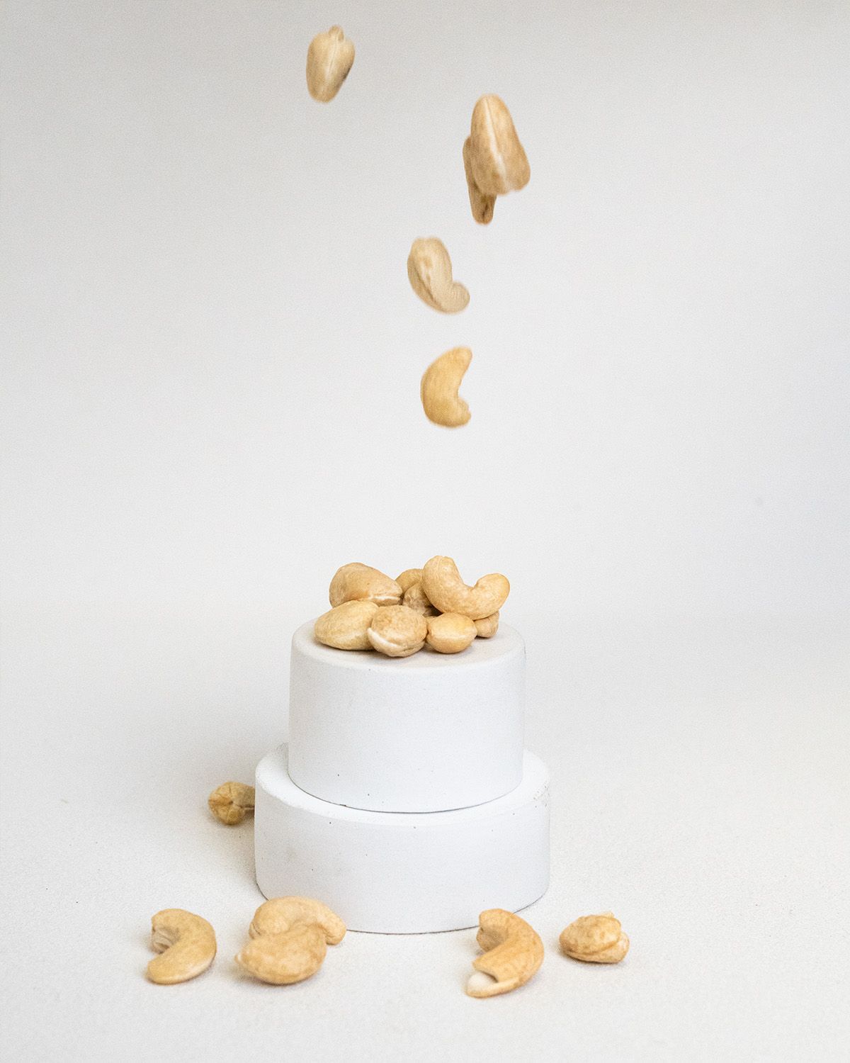 Cashewkerne fallen auf einen weißen Sockel. Einige Nüsse liegen verstreut. Helle, beige Farbe.