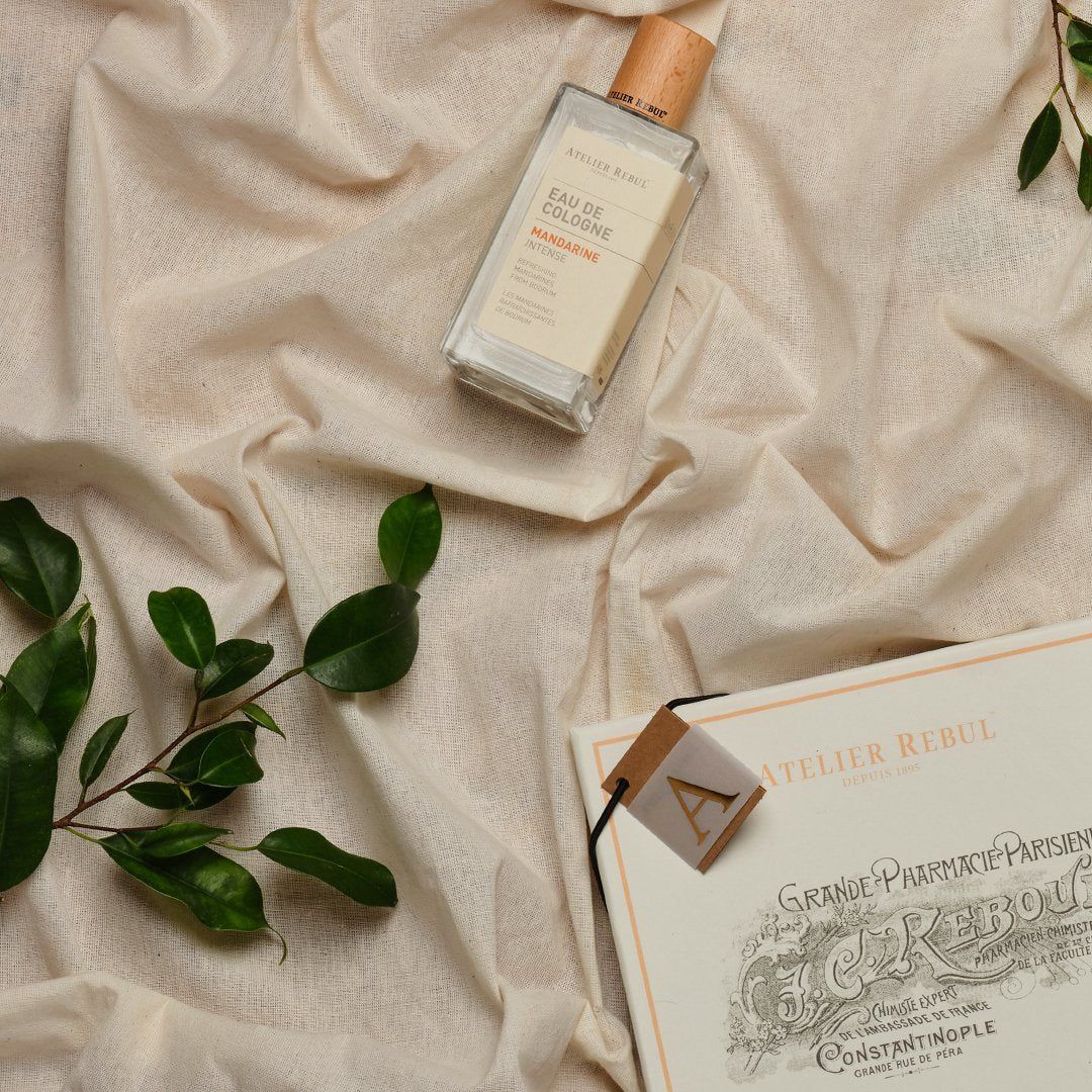 Glasflasche Eau de Cologne mit Holzverschluss. Aufschrift: Atelier Rebul Mandarine Eau de Cologne. Auf cremefarbenem Stoff.