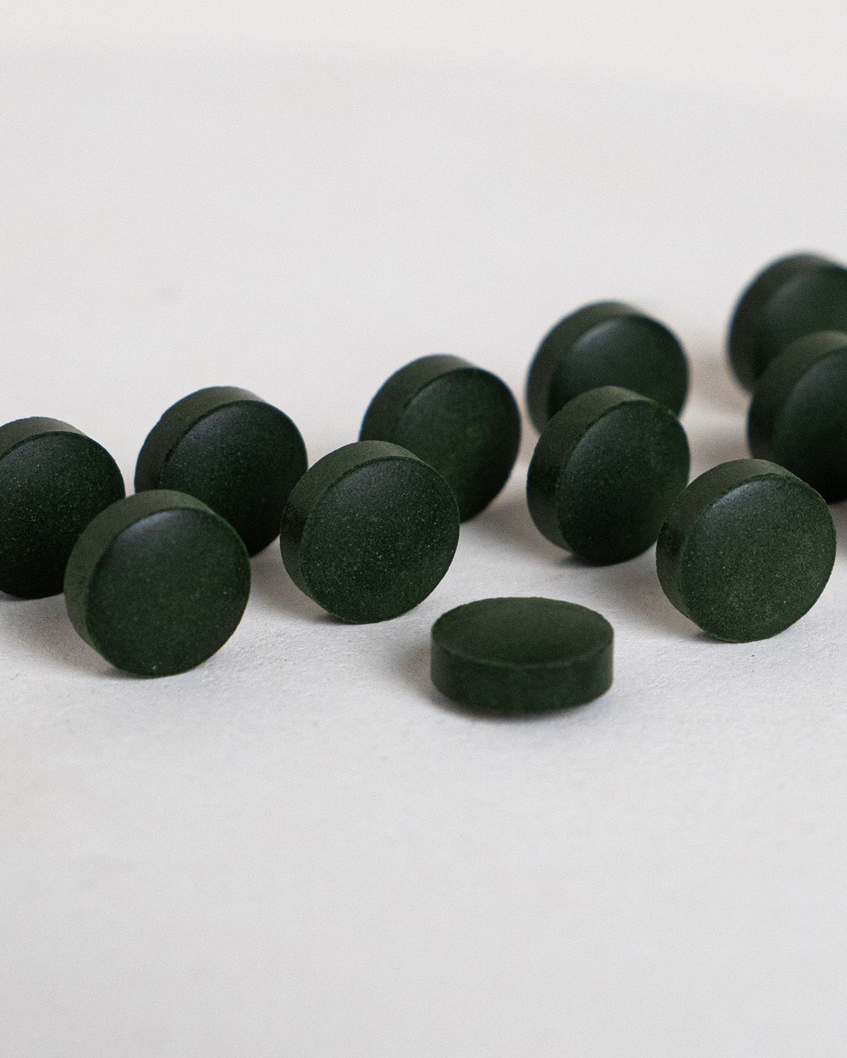 Dunkelgrüne Tabletten, rund und flach. Einige Tabletten liegen verstreut auf weißem Untergrund.