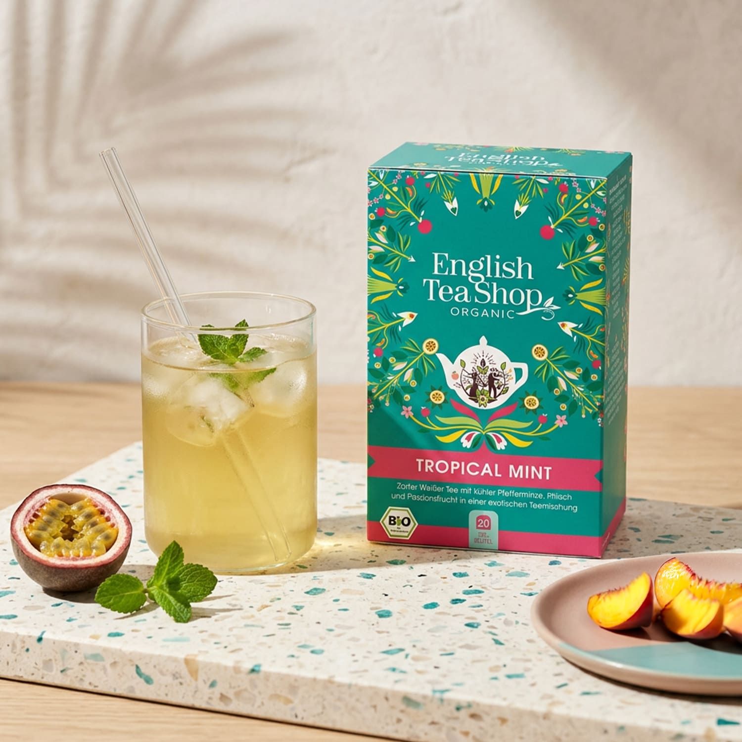 Glas mit Eis, Tee und Minze. Daneben Teebox "English Tea Shop Organic Tropical Mint". Pfirsich und Passionsfrucht auf Platte.