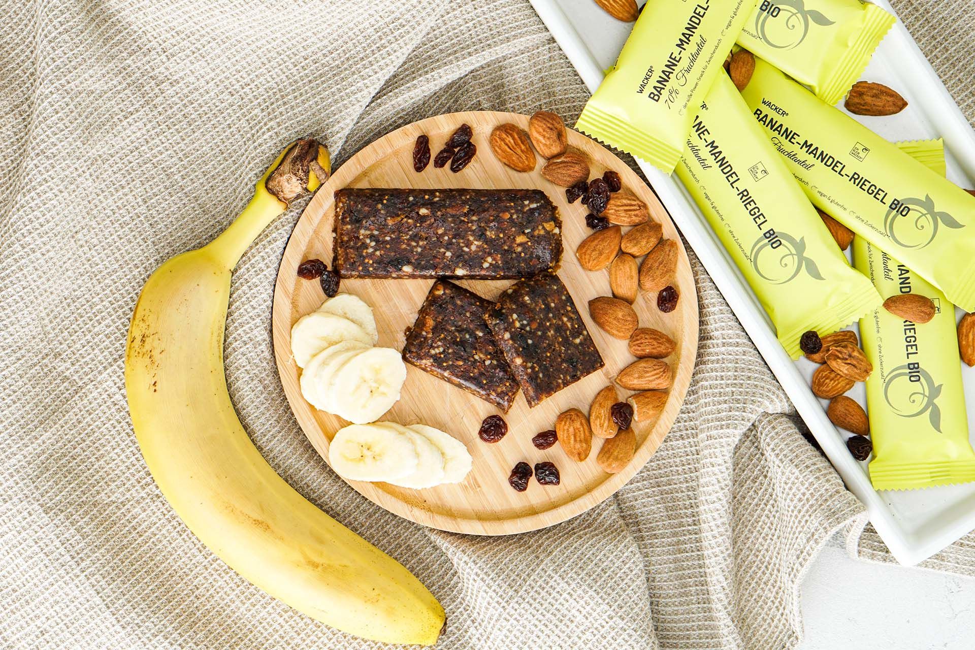 Banane, geschnittene Riegel, Mandeln, Rosinen auf Holzplatte. Gelbe Riegel im Hintergrund.