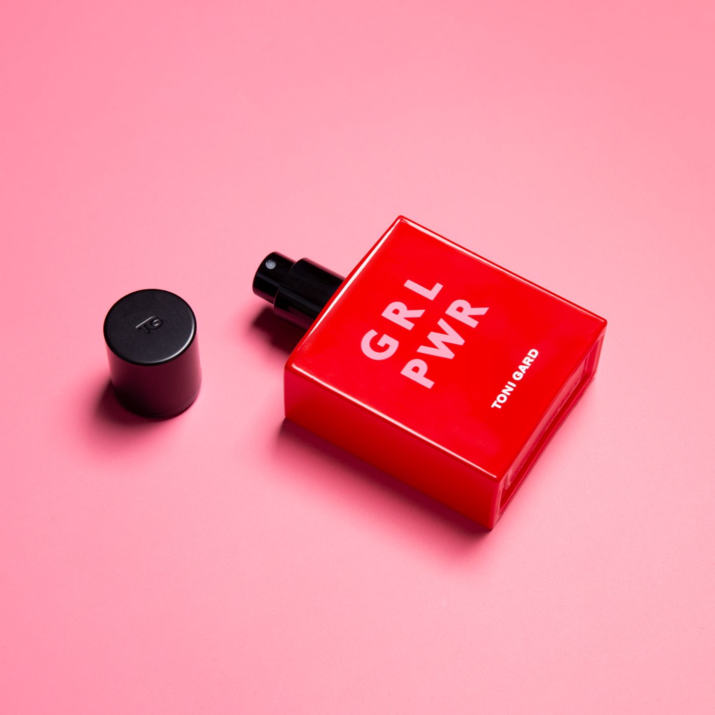 Rote rechteckige Flasche mit schwarzem Verschluss. Text: GRL PWR, TONI GARD. Schwarzer Deckel daneben. Rosa Hintergrund.