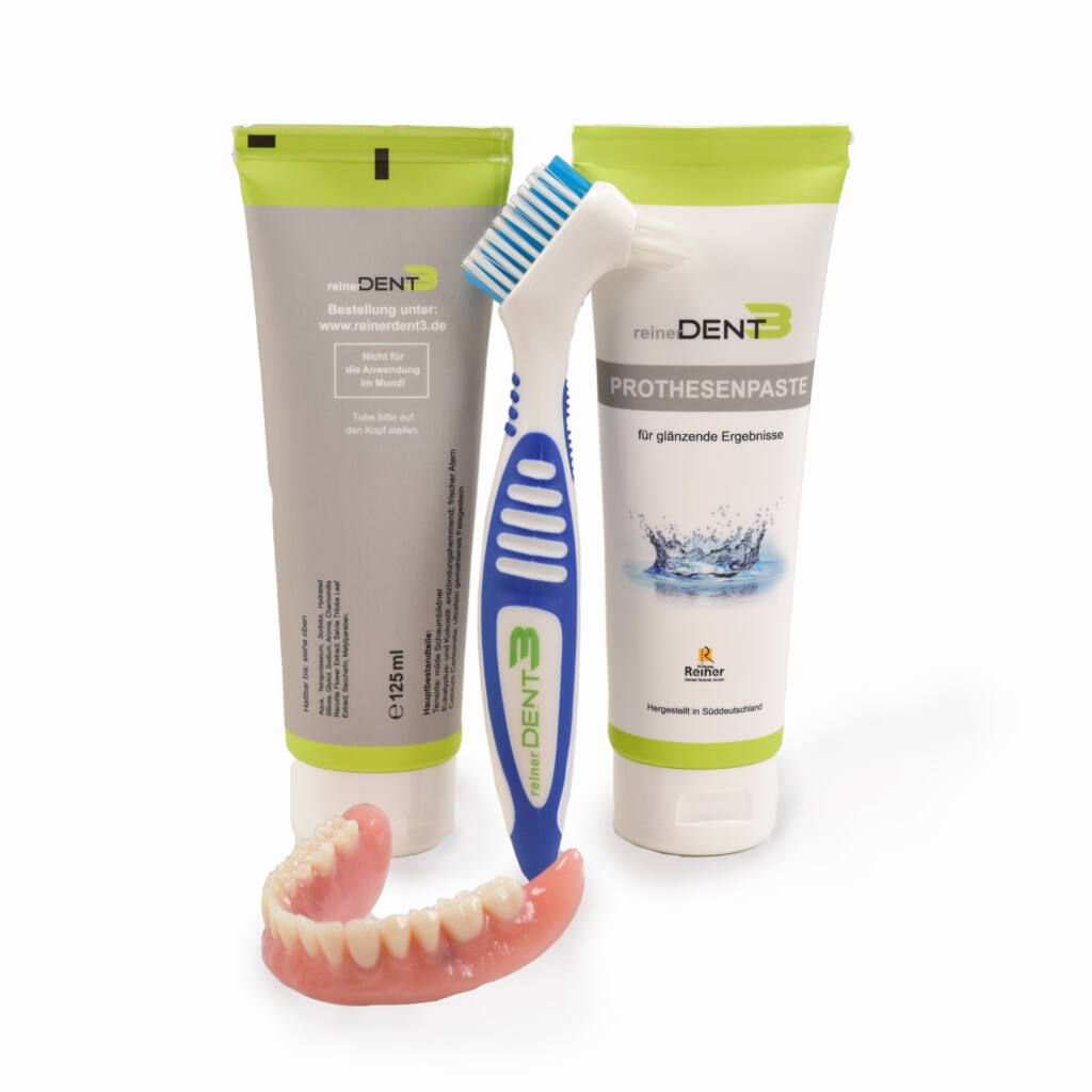 Zwei Tuben Zahnprothesenpaste, eine Zahnbürste und eine Zahnprothese. Tuben mit Produktnamen. Zahnbürste blau-weiß. Prothese rosa.