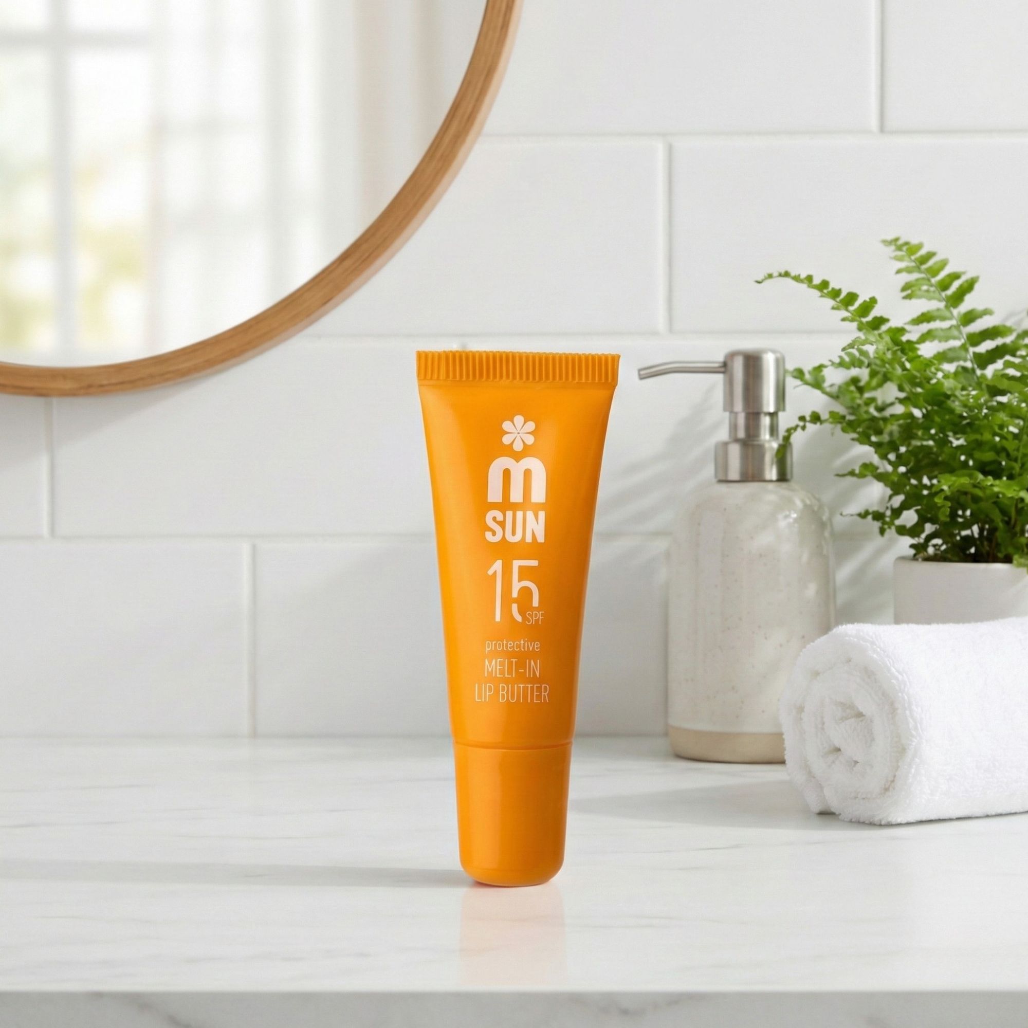 Orange Lippenbutter-Tube auf weißer Oberfläche. Aufschrift: SUN 15 SPF, protective MELT-IN LIP BUTTER. Im Hintergrund Spiegel, Seife, Handtuch.