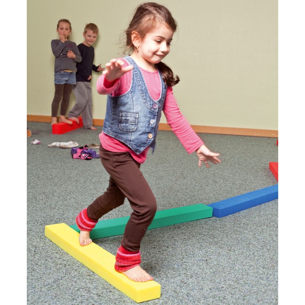 Kind balanciert auf gelben Schaumstoffbausteinen. Weitere farbige Bausteine liegen auf dem Boden. Im Hintergrund stehen Kinder.