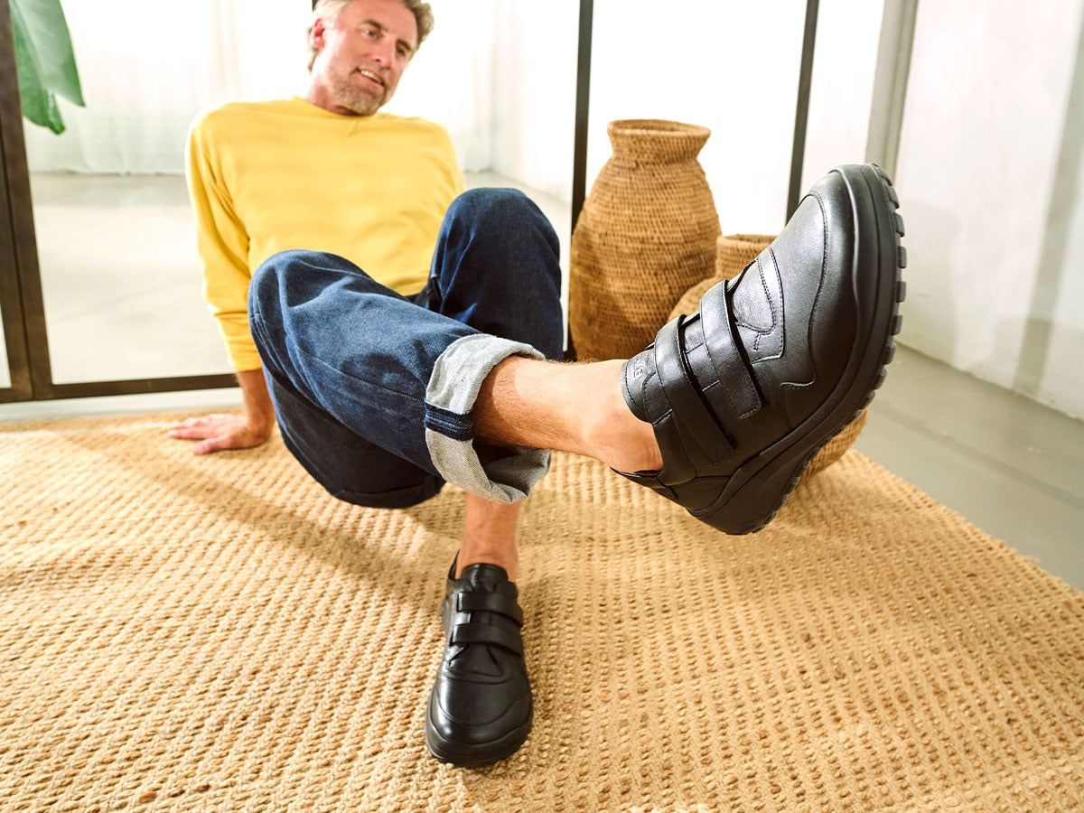 Mann sitzt auf dem Boden, trägt schwarze Herren-Slipper mit Klettverschluss. Breite Passform. Leder. Auf Teppich.