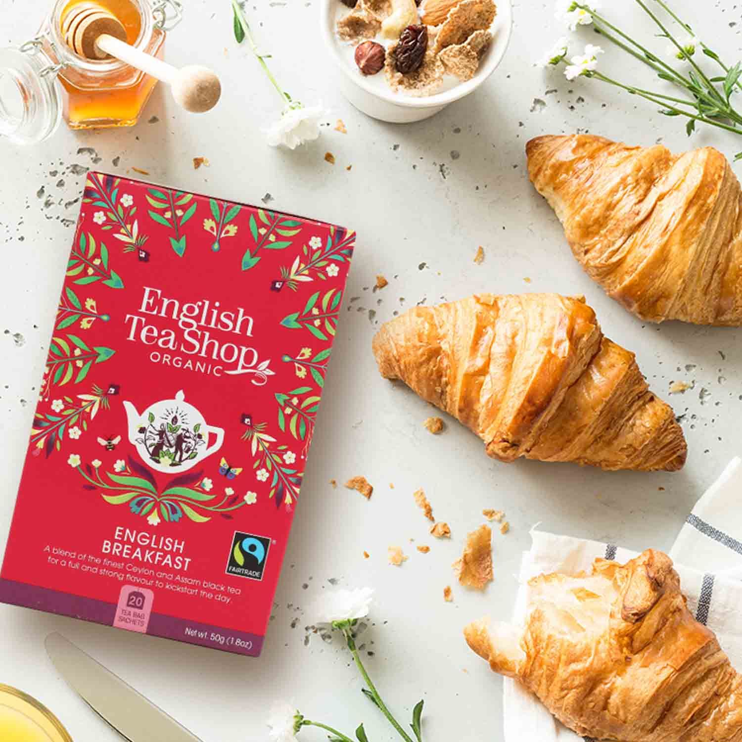 Rote Schachtel mit Aufschrift "English Tea Shop Organic English Breakfast". Daneben Croissants, Honig und Müsli.