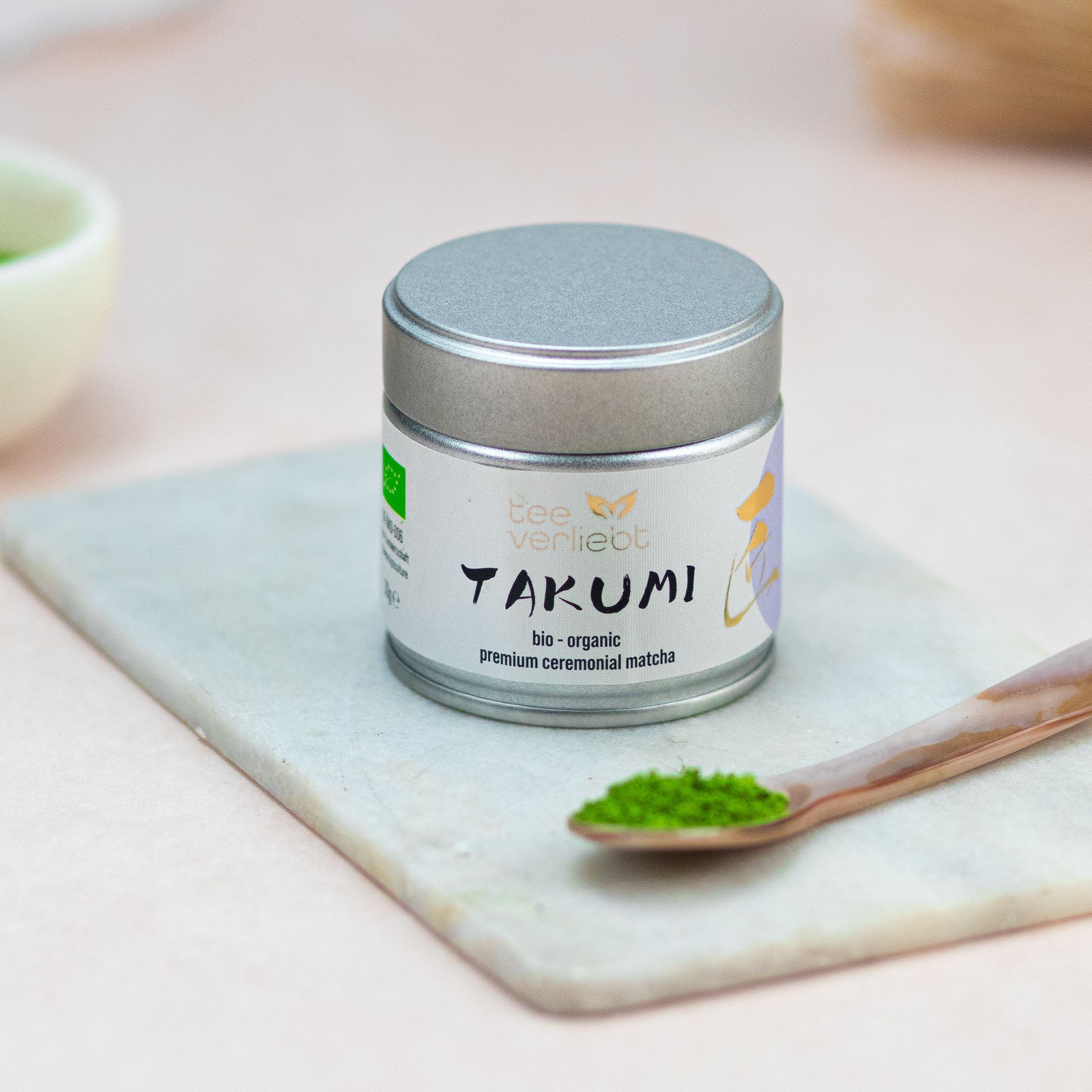 Dose auf Marmorplatte mit Löffel Matcha. Aufschrift: teeverliebt, TAKUMI, bio-organic, premium ceremonial matcha. Bio-Siegel.