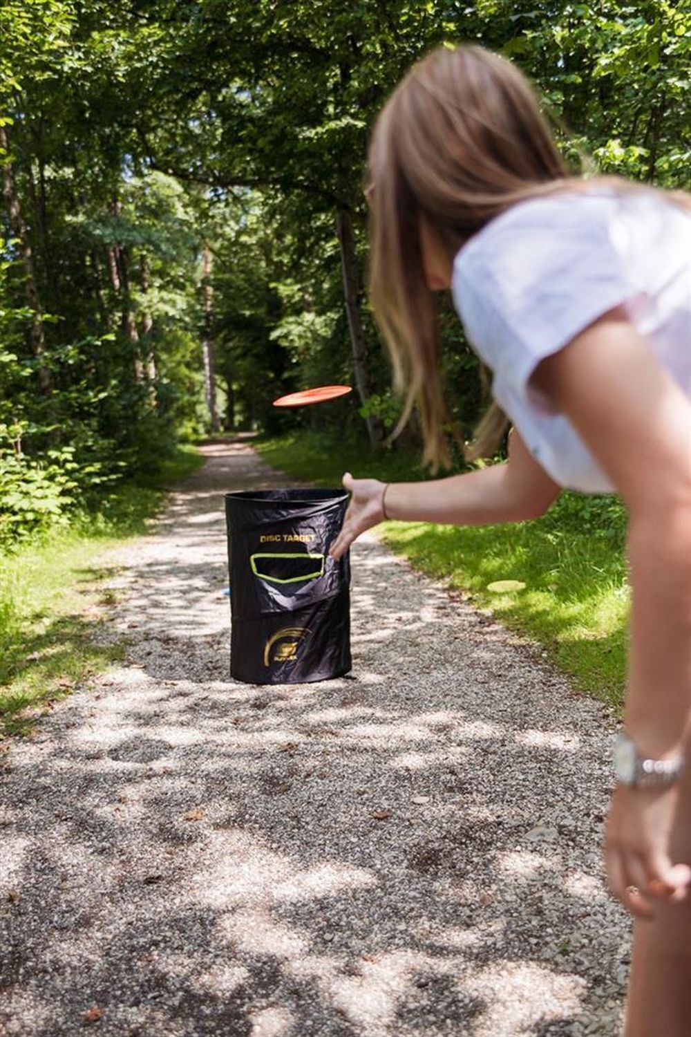 Person wirft Frisbee auf Disc Golf Ziel. Schwarzes Ziel mit gelbem Schriftzug "DISC TARGET". Auf einem Weg im Wald.