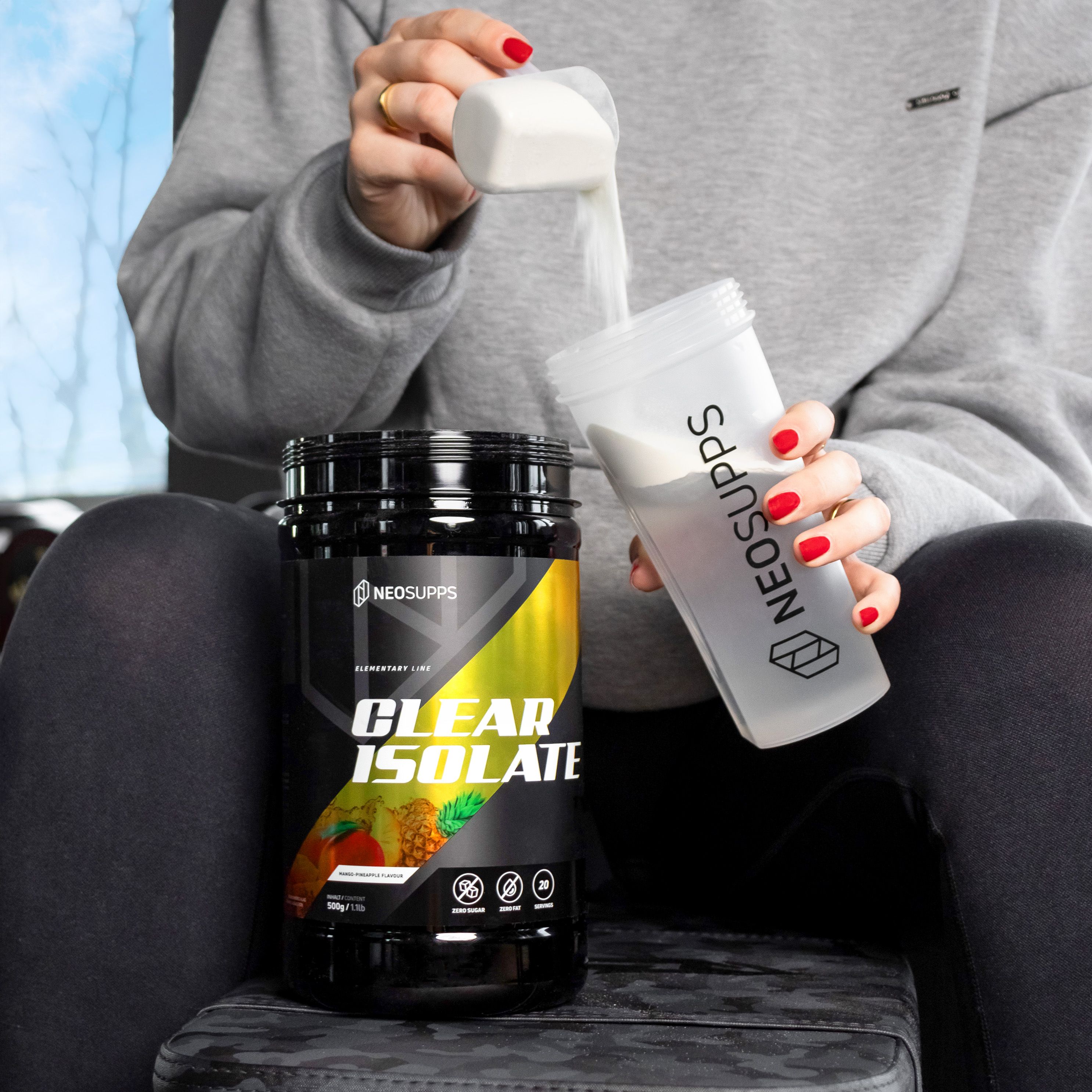 Person gießt Pulver aus Dose "CLEAR ISOLATE" in Shaker. Dose und Shaker mit Logo. Rote Fingernägel.
