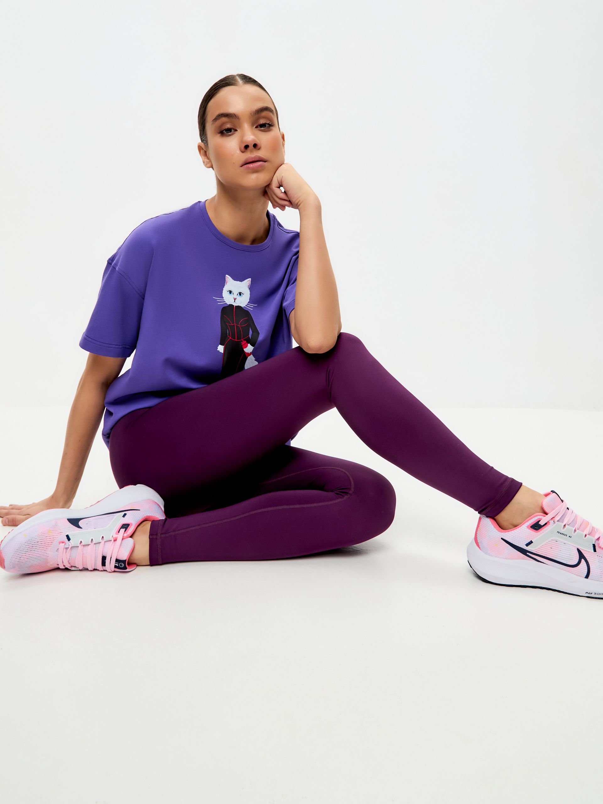 Dunkelviolette Leggings. Frau trägt lila T-Shirt mit Katzenmotiv. Pinke Schuhe. Sitzende Pose.