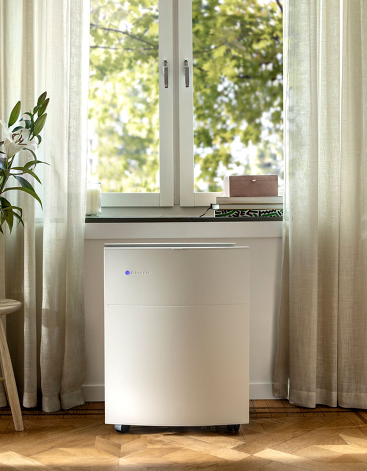 Luftreiniger vor Fenster. Weißes Gehäuse, Rollen. Steht auf Holzboden. Vorhänge im Hintergrund.