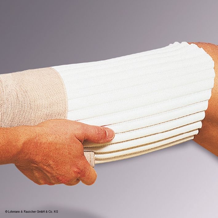 Komprex® Schaumstoff Platte II, weiß, gerippt, am Arm angebracht. Beige Bandage fixiert das Produkt. Logo: Lohmann & Rauscher.