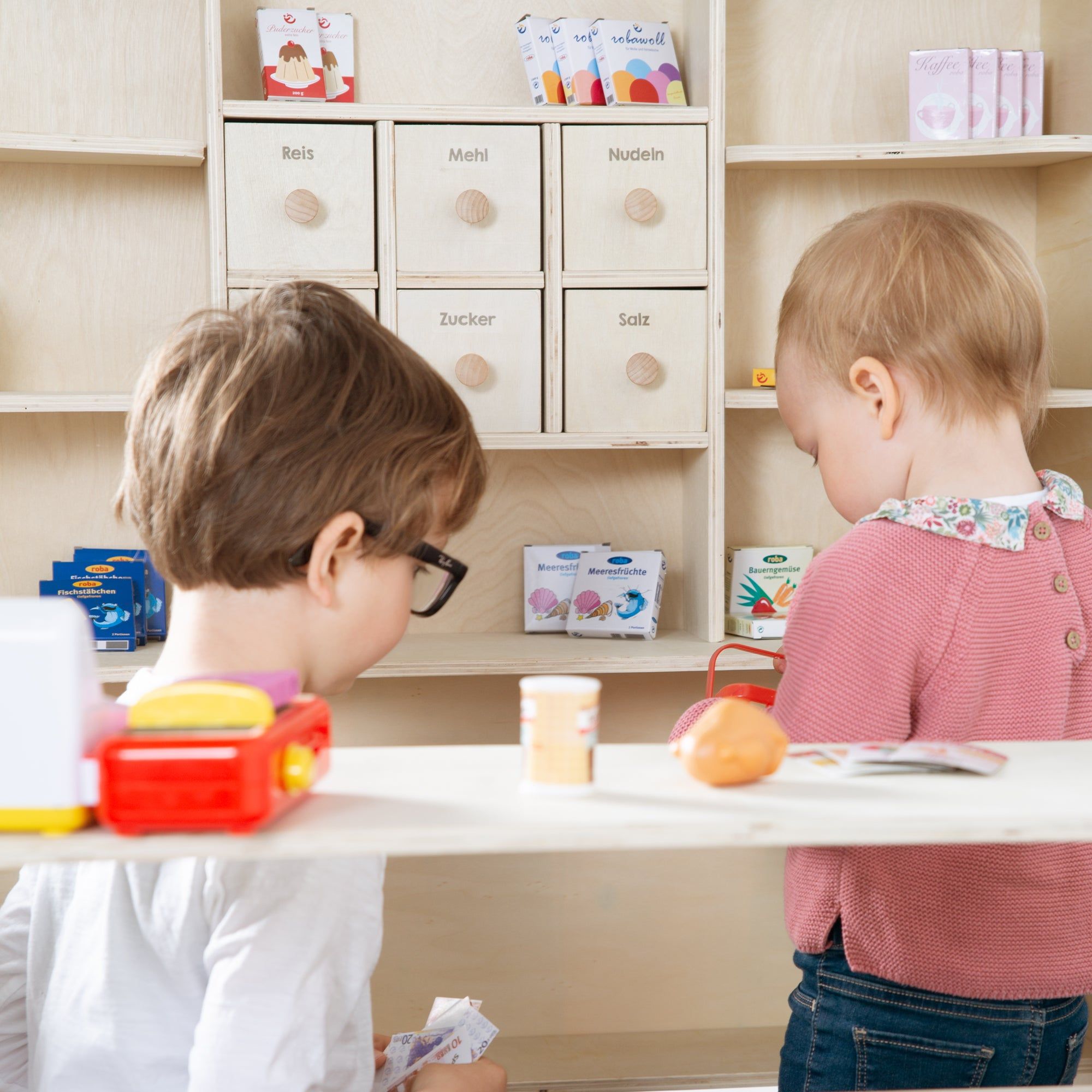 Kinder spielen an einem Holz-Verkaufsstand. Regale mit Schubladen und Spielzeug-Lebensmitteln. Aufschrift: Little Shop.
