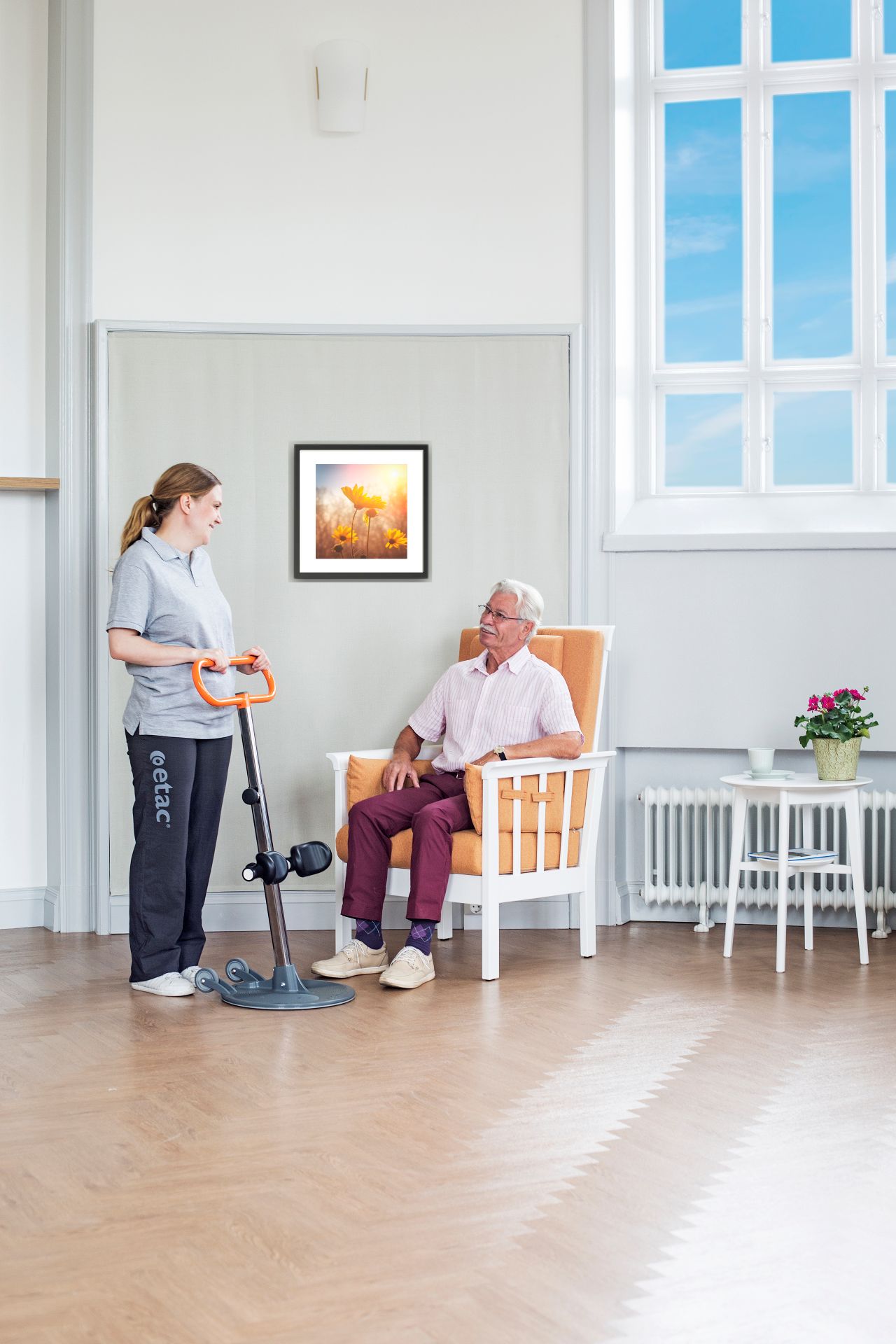 Pflegekraft mit Transferhilfe neben sitzendem Mann. Zimmer mit Fenster, Bild und Sessel.