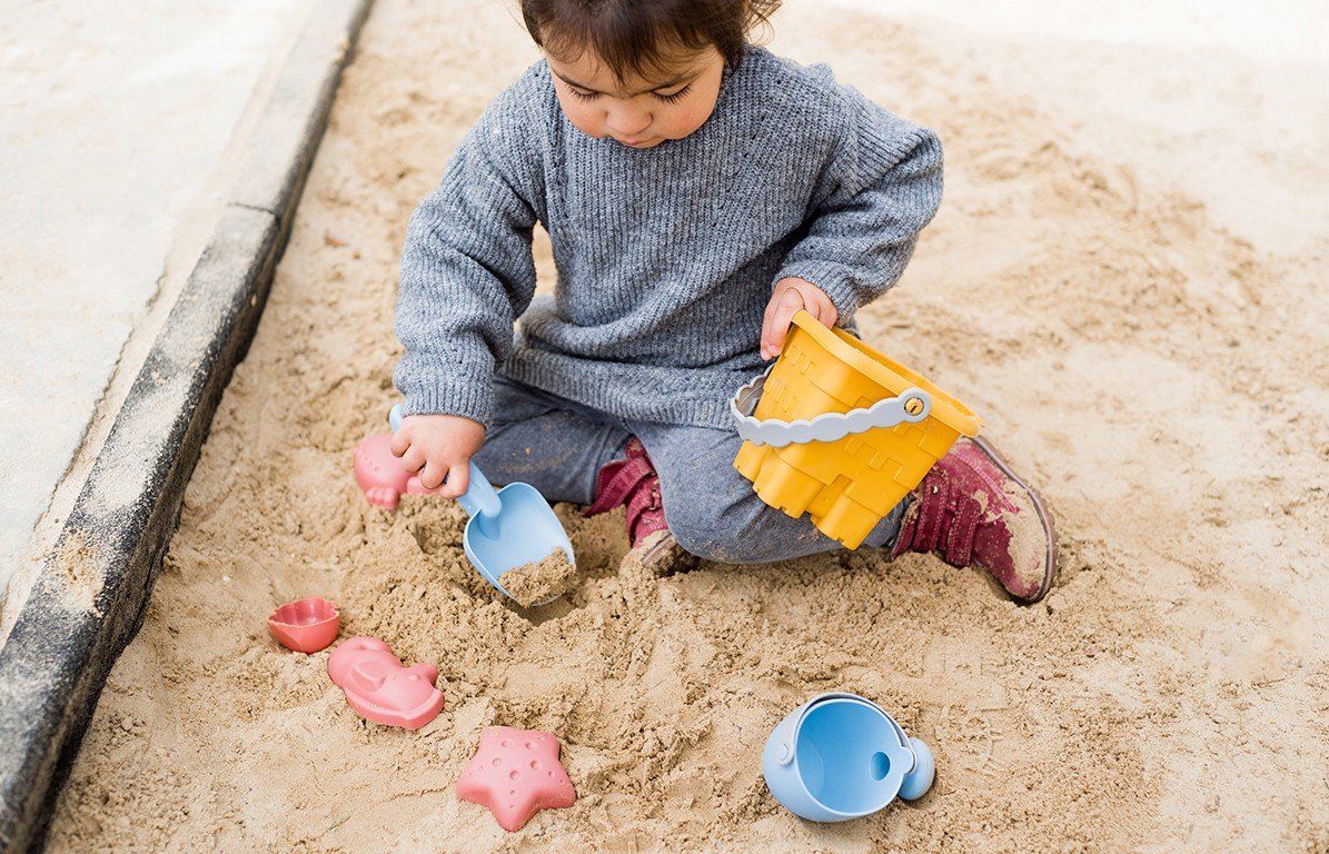 Kind spielt im Sandkasten mit gelbem Eimer, blauer Schaufel und rosa Formen. Sandspielzeug auf Sand.