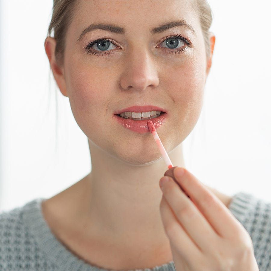 Frau trägt GRN Lipgloss auf. Sie hält den Applikator an ihren Lippen. Helle Haut, grauer Pullover.