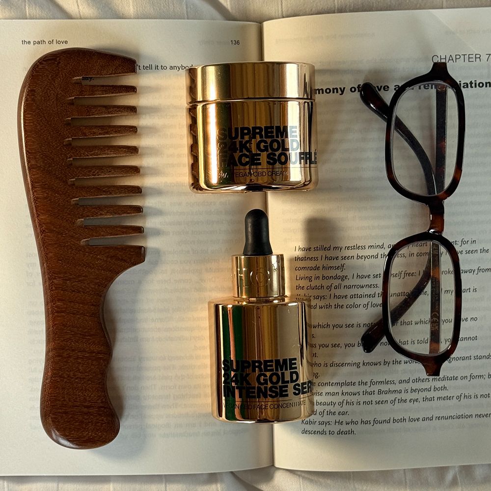 Goldene Serumflasche und Creme auf Buch. Aufschrift: Supreme 24K Gold Intense Serum. Mit Kamm und Brille.